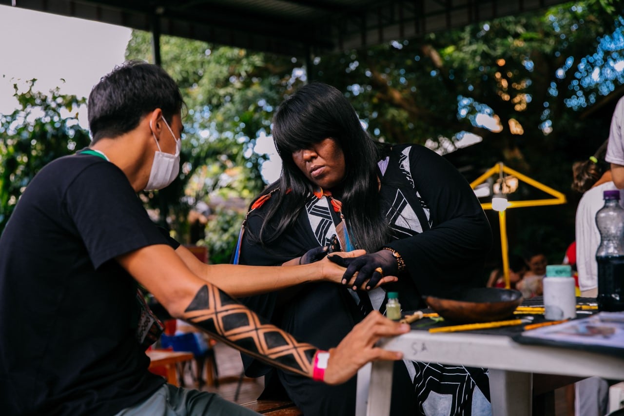 Mulher indígena fazendo pintura em homem durante a Feira da FAS, realizada pela Fundação Amazônia Sustentável (FAS).