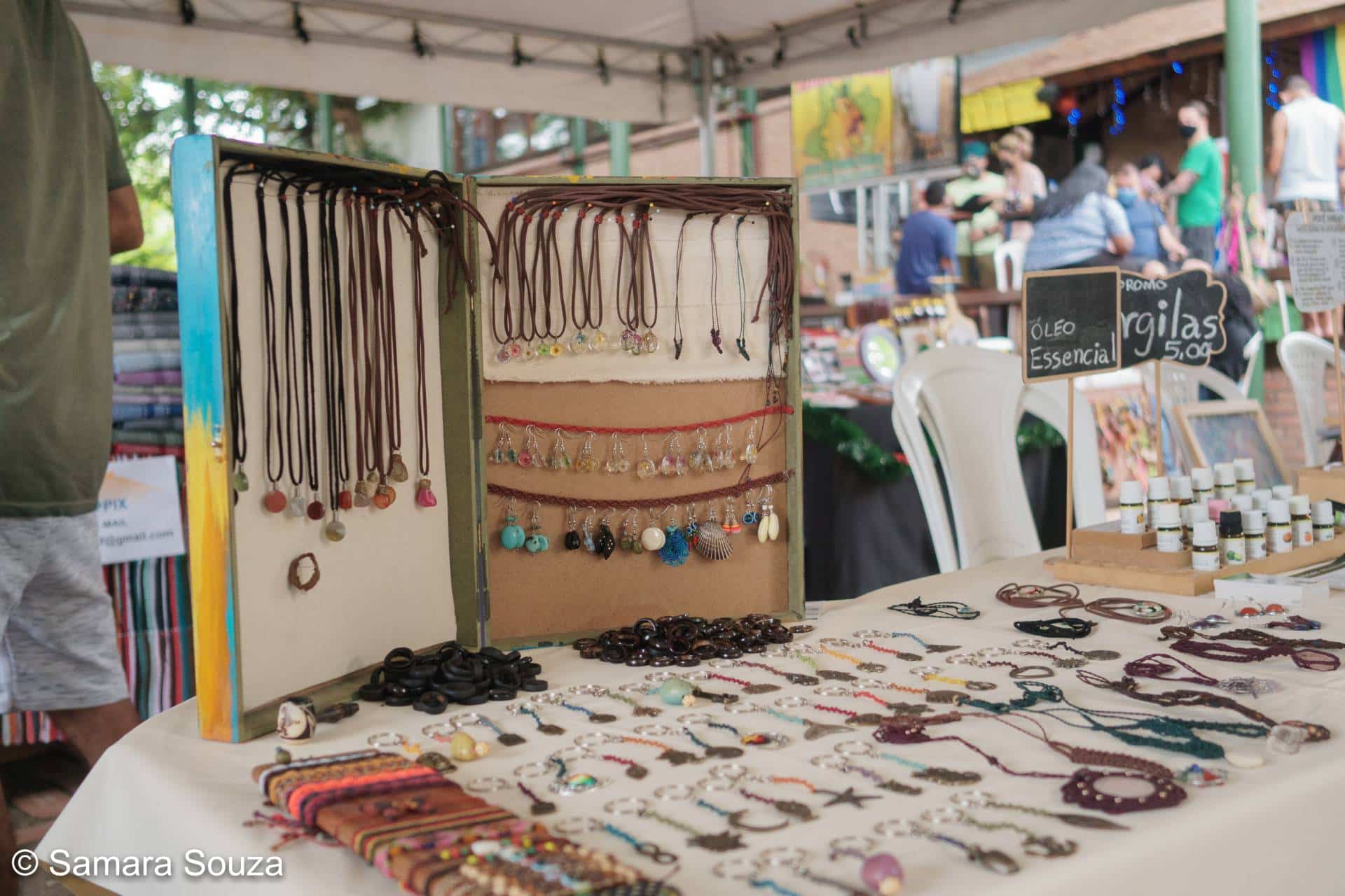Vários artesanatos expostos em uma mesa, incluindo pulseiras, cordões, brincos e anéis.
